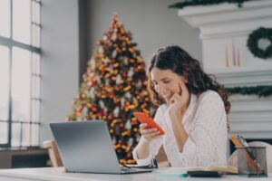 woman texting and on computer with christmas tree in background